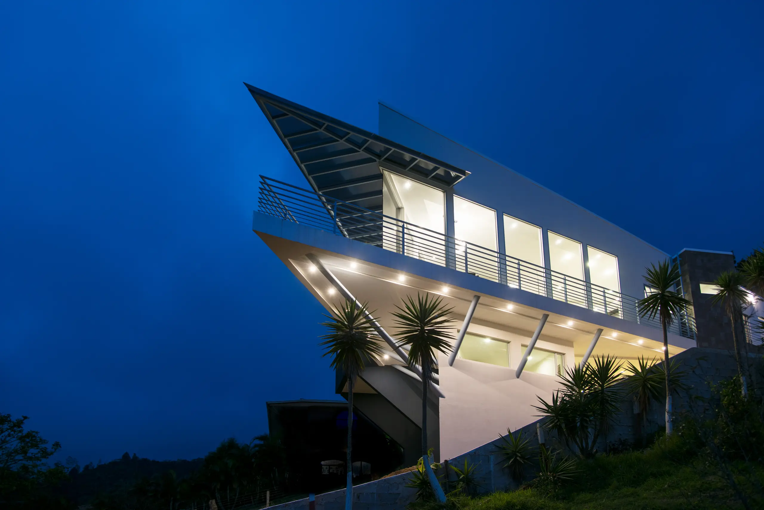 Casa blanca de noche con terraza en voladizo sobre puntales inclinados, alero de cristal y ventanales encendidos