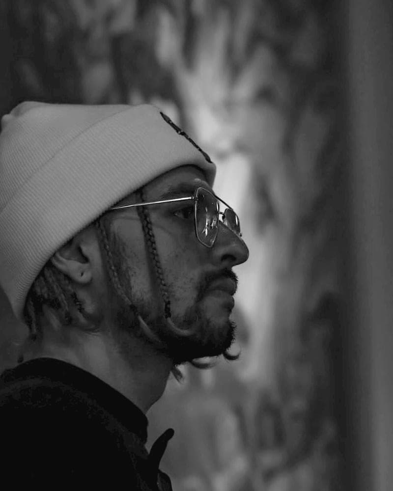 Hombre de perfil con gorro tejido, anteojos y barba, en penumbra frente a un mural