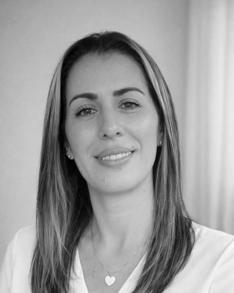 Mujer de cabello largo y blusa blanca, sonriendo de frente ante un fondo claro y liso