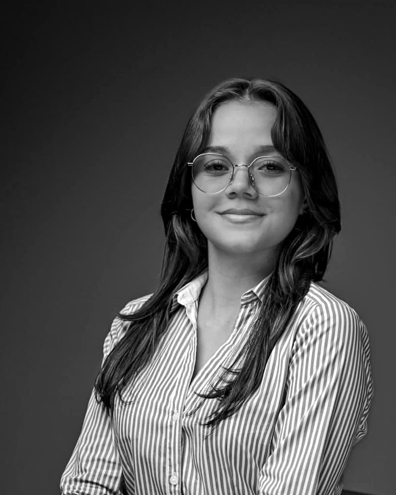 Joven con anteojos redondos y camisa a rayas, sentada y sonriendo ante un fondo oscuro de estudio