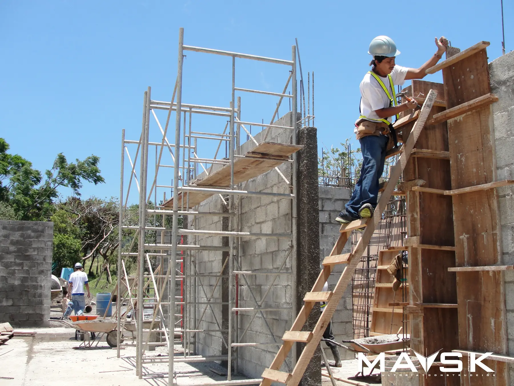 Obrero con casco subido a una escalera, armando el encofrado de madera de una columna junto a los andamios